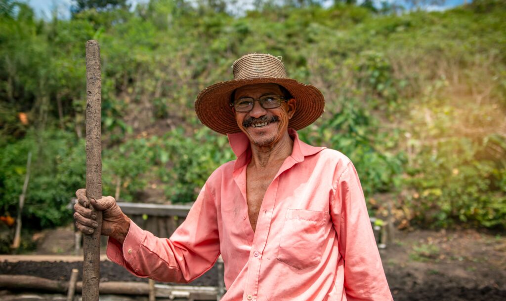 Homem idoso em área rural. Ele veste uma camisa rosa. Segura um cajado e está de chapéu de palha. Ele está perto de um barranco de rio. 

Ele ilustra o capítulo sobre como dar entrada na aposentadoria rural. 